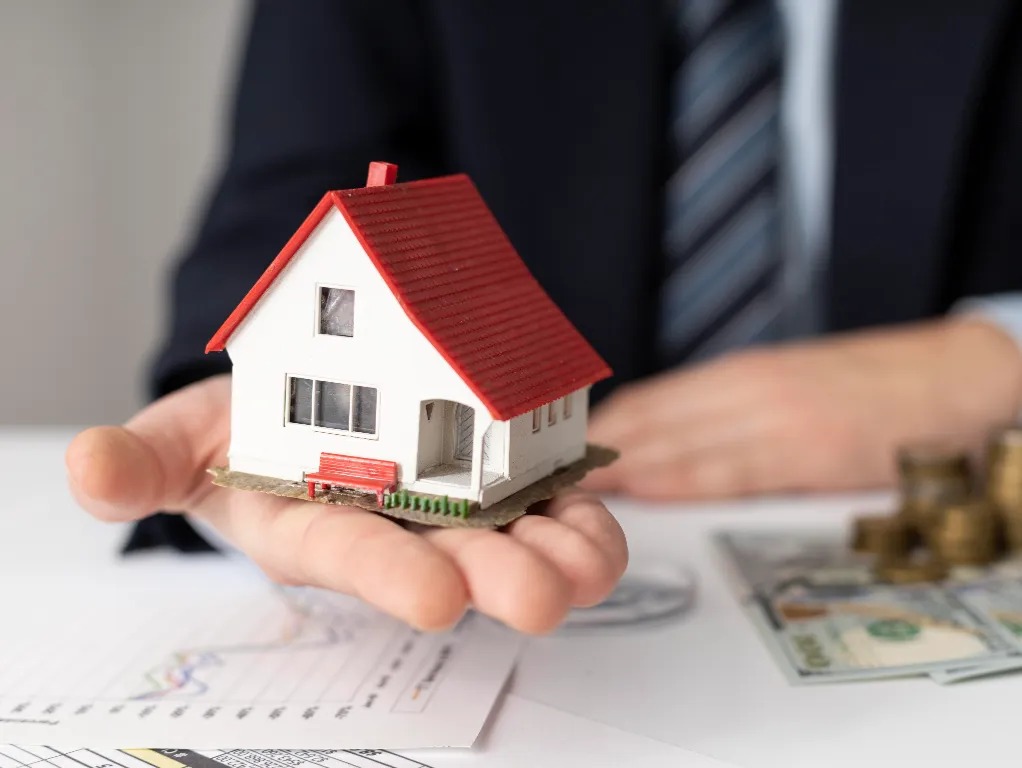 A person in a business suit holding a small house model over financial documents, symbolizing REACH Housing Solutions’ mission to expand affordable housing and community partnerships for individuals living with mental health challenges.