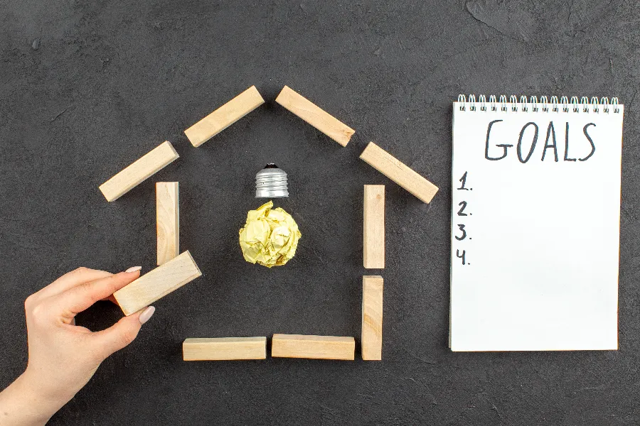 Wooden blocks forming a house with a light bulb inside and a notepad labeled “Goals,” symbolizing Reach Housing Solutions’ mission to create supportive community-based housing and empower residents to achieve recovery and stability.